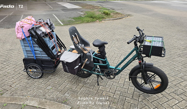 Se muestra una bicicleta eléctrica de carga Fiido T2 completamente cargada, con un asiento infantil, alforjas laterales, una caja de carga delantera y un remolque trasero muy cargado.