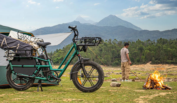 Bicicleta eléctrica de carga Fiido T2 cargada con equipo de camping en un campamento de montaña, junto a una tienda de campaña y una fogata